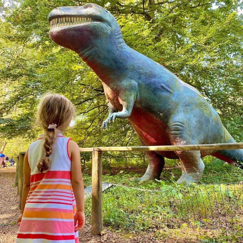 A young child enjoying the dinosaur trail - one of many things for families to enjoy at Knebworth this autumn. She's wearing a white top, a pink striped skirt and is looking up at a model of a T-Rex.
